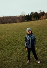 Enfant portant une veste camo bleu marine et grise, un pantalon noir et un bonnet gris, se tenant sur l'herbe. Des baskets beiges avec des lacets complètent la tenue.