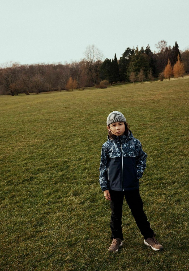 Enfant portant une veste camo bleu marine et grise, un pantalon noir et un bonnet gris, se tenant sur l'herbe. Des baskets beiges avec des lacets complètent la tenue.