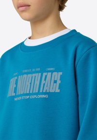 Turquoise sweatshirt with ribbed crew neck, featuring bold white print "THE NORTH FACE" and "NEVER STOP EXPLORING" in a geometric font.
