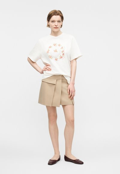 Ung kvinne med kort hår iført hvit t-skjorte med blomstermønster i rundform, beige plissert miniskjørt og mørkebrune flate sko, stående med hånden på hoften.