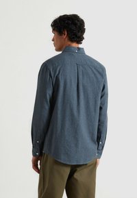 Ciemnoniebieska koszula z długim rękawem zapinana na guziki, z detalem w postaci plis na plecach i zaokrąglonym dołem; zestawiona z oliwkowymi spodniami.