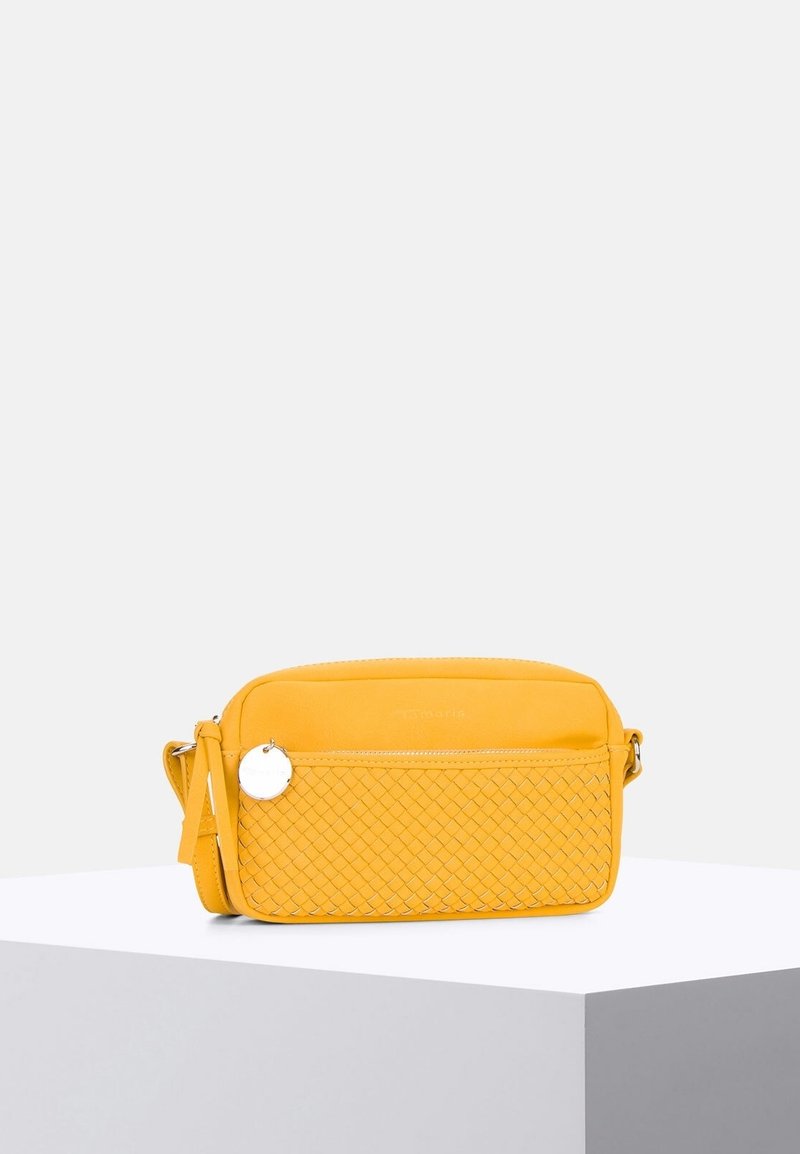 Borsa a tracolla gialla con tasca anteriore intrecciata, ciondolo rotondo in argento e tracolla regolabile su un piedistallo bianco.