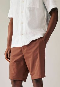 Shorts marron au toucher lisse, dotés de poches latérales. Associés à une chemise en lin blanche à manches courtes avec une poche poitrine et des boutons.