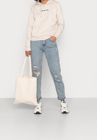 Persona con felpa leggera beige, jeans chiari strappati, sneakers bianche Adidas, che tiene una borsa tote beige semplice, in piedi davanti a uno sfondo neutro.