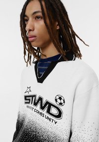Pull blanc et noir avec un col en V, présentant un logo graphique et des accents de ballon de football. Le tissu a un motif texturé et en dégradé.