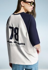 Bela in mornarsko modra barvno blokirana kratka majica s kratkimi rokavi, ki na hrbtni strani prikazuje velike črne številke "78" in besedilo "ANGEL NUMBERS". Ohlapna zasnova, mehak material.