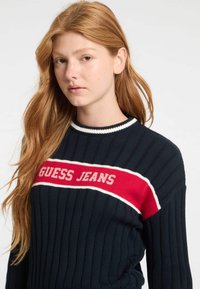 Marinblå ribbad tröja med ett rödvitt randigt band som har "GUESS JEANS" i vit text. Stickad struktur, rund halsringning, långa ärmar.