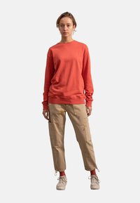 Felpa arancione in cotone con maniche lunghe, abbinata a pantaloni cargo beige con tasche e dettagli di vernice. Indossati con scarpe chiare.