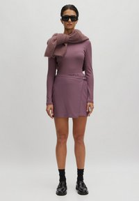 Paarse geribde lange mouw top gecombineerd met een wikkelrok en een bijpassende sjaal. Glanzende zwarte schoenen en zwarte sokken maken de outfit compleet.
