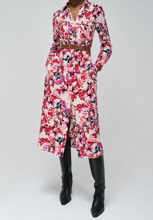 Kobieta ubrana w długą, zapinaną na guziki sukienkę w kwiaty z czerwonymi, różowymi i zielonymi wzorami, brązowy pasek oraz czarne kozaki za kolano.