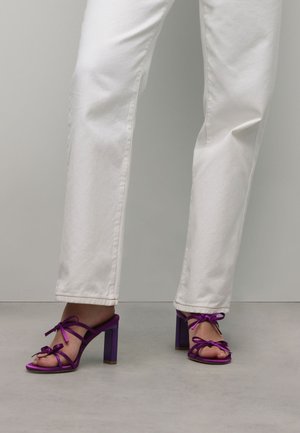 Purple satin high-heeled sandals with dual bows, open toe, and a square block heel. Worn with white straight-leg jeans.