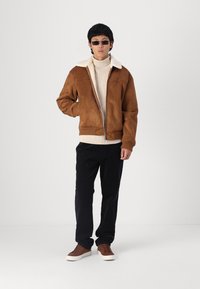 Rudos suede bomber tipo švarkas su balta avikailio apykakle, kreminės spalvos golfas, juodos kelnės ir rudi sportbačiai. Modelis stovi su viena ranka kišenėje.