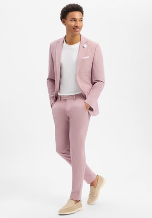 Mann trägt einen hellrosa Anzug mit weißem T-Shirt und beigen Slippern, steht mit den Händen in den Taschen, lächelt und schaut zur Seite.