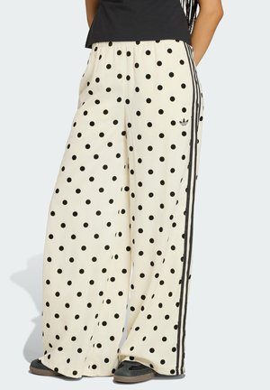 Bela široke hlače s črnimi pikami, črnimi Adidas črtami na straneh in elastičnim pasom, nošene s črnimi čevlji in črnim zgornjim delom.