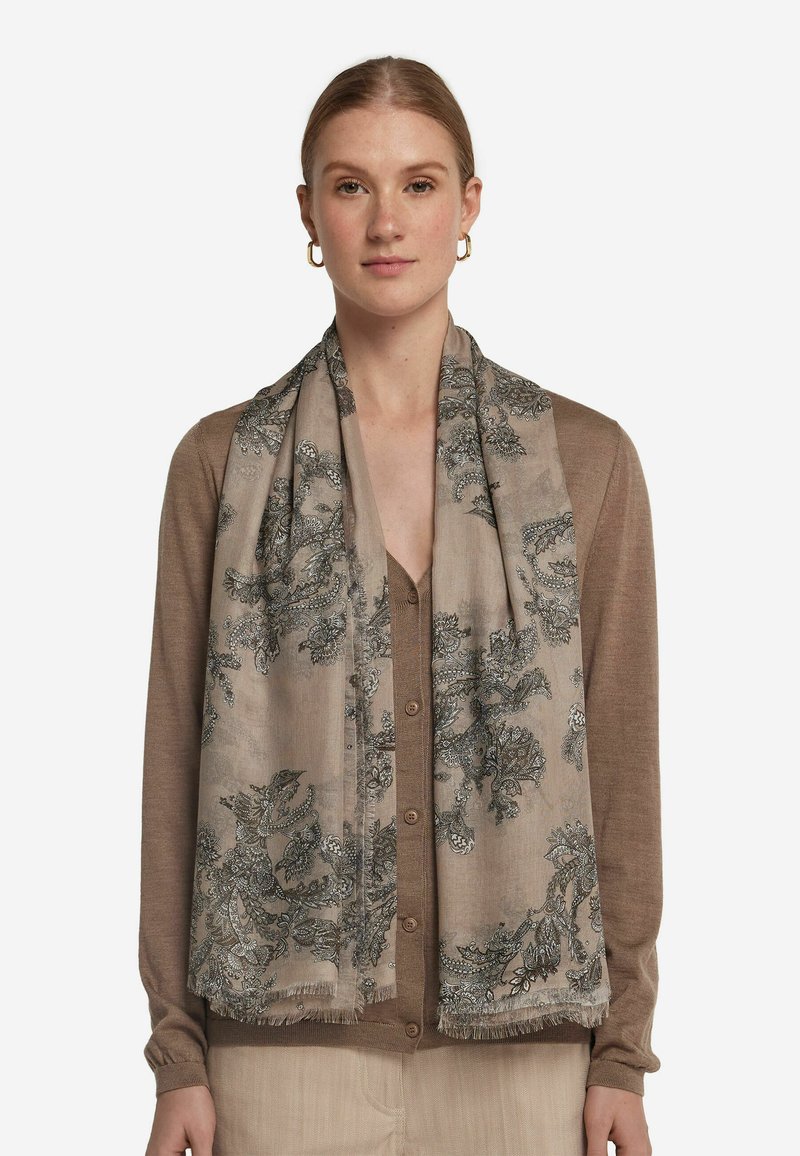 Écharpe légère beige avec des motifs floraux sombres complexes, aux bords frangés, drapée élégamment sur un cardigan brun.