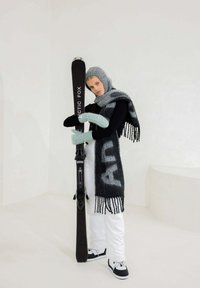 Ski noir avec le texte "ARCTIC FOX", écharpe à franges grise, gants en tricot bleu clair, pantalon blanc, sneakers noirs et fond neutre.
