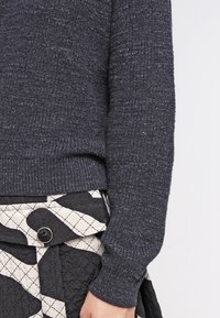 Manche de pull en tricot gris foncé et jupe matelassée noire et blanche avec une poche à rabat boutonnée visible sur le côté.