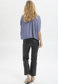 Femme aux longs cheveux blonds portant un haut violet ample, un pantalon noir ajusté et des sandales ouvertes de couleur claire, se tenant contre un fond blanc.