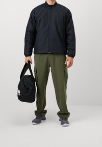 Svart vadderad jacka med dragkedja, gröna cargo-byxor med sidofickor och grå sportskor. Svart duffelväska med en vit logotyp.