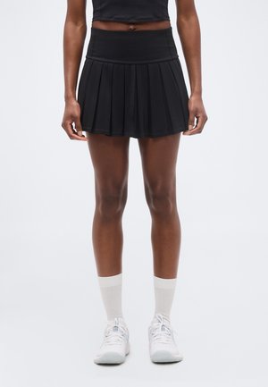 Γυναίκα που φοράει μαύρη πιετέ αθλητική φούστα, λευκές κάλτσες μέχρι τον αστράγαλο και λευκά αθλητικά παπούτσια, στέκεται μπροστά σε απλό φωτεινό φόντο.