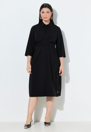 Kobieta w czarnej sukience midi z rękawami do 3/4, kołnierzykiem i zawiązywanym paskiem w talii, w połączeniu z czarnymi szpilkami z ostrym noskiem.