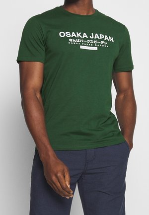 Mies, jolla on tummanvihreä T-paita, jossa on valkoinen teksti "OSAKA JAPAN Namba Parks Garden", ja tummansiniset housut, seisoo harmaata taustaa vasten.