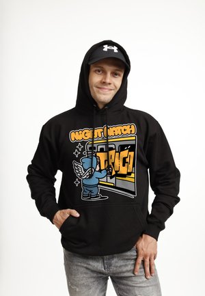 Joven sonriendo con sudadera negra con arte de graffiti de dibujos animados y gorra negra, de pie contra un fondo blanco.