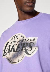 Tricou din bumbac mov, cu un logo "Los Angeles Lakers" în culorile negru și aur, însoțit de o grafică cu baschet. Gât rotund, mâneci scurte.