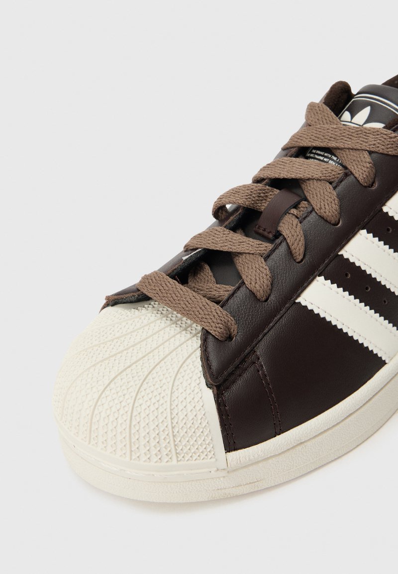 Primo piano di una scarpa da ginnastica in pelle marrone con punta beige a guscio, suola in gomma beige, lacci marroni e le iconiche tre strisce bianche Adidas sul lato.