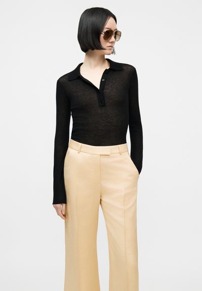 Kvinne iført svart, langermet, gjennomsiktig poloshirt, beige høytaljede vide bukser og store brune solbriller, stående med én hånd i lommen.