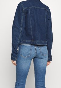 Tummansininen farkkutakki, jossa on tiukka muotoilu, näkyvät saumat ja lyhennetty helma, yhdistettynä vaaleansinisiin slim fit -farkkuihin, joissa on takataskut.