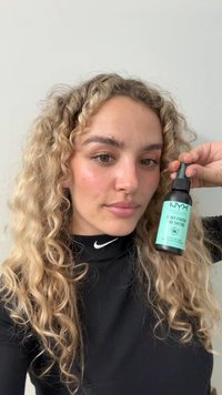Bouteille en spray noire avec une étiquette turquoise indiquant "Fini Rosé" tenue par une personne aux cheveux bouclés. Pas de motifs ou de logos visibles sur les vêtements.