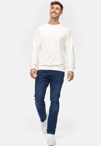 Hvit hettegenser med ribbestrikkede mansjetter og kant, kombinert med mørkeblå jeans og hvite joggesko. Enkel design uten synlige logoer eller mønstre.