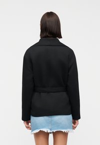 Femme aux cheveux bruns raides portant une veste noire ceinturée et une mini-jupe en denim bleu clair effilochée, vue de dos sur un fond blanc.