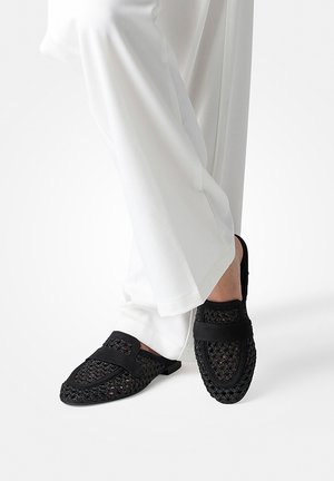 Schwarze gewebte Slip-On-Loafer, getragen mit weißen weiten Hosen, die einen entspannten und stylischen Casual-Look zeigen.