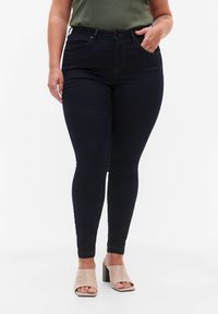 Vaqueros skinny de color azul oscuro con una cintura alta, ligera elasticidad, con bolsillos estándar y costuras doradas en contraste. Combinados con sandalias de tacón cuadrado beige.