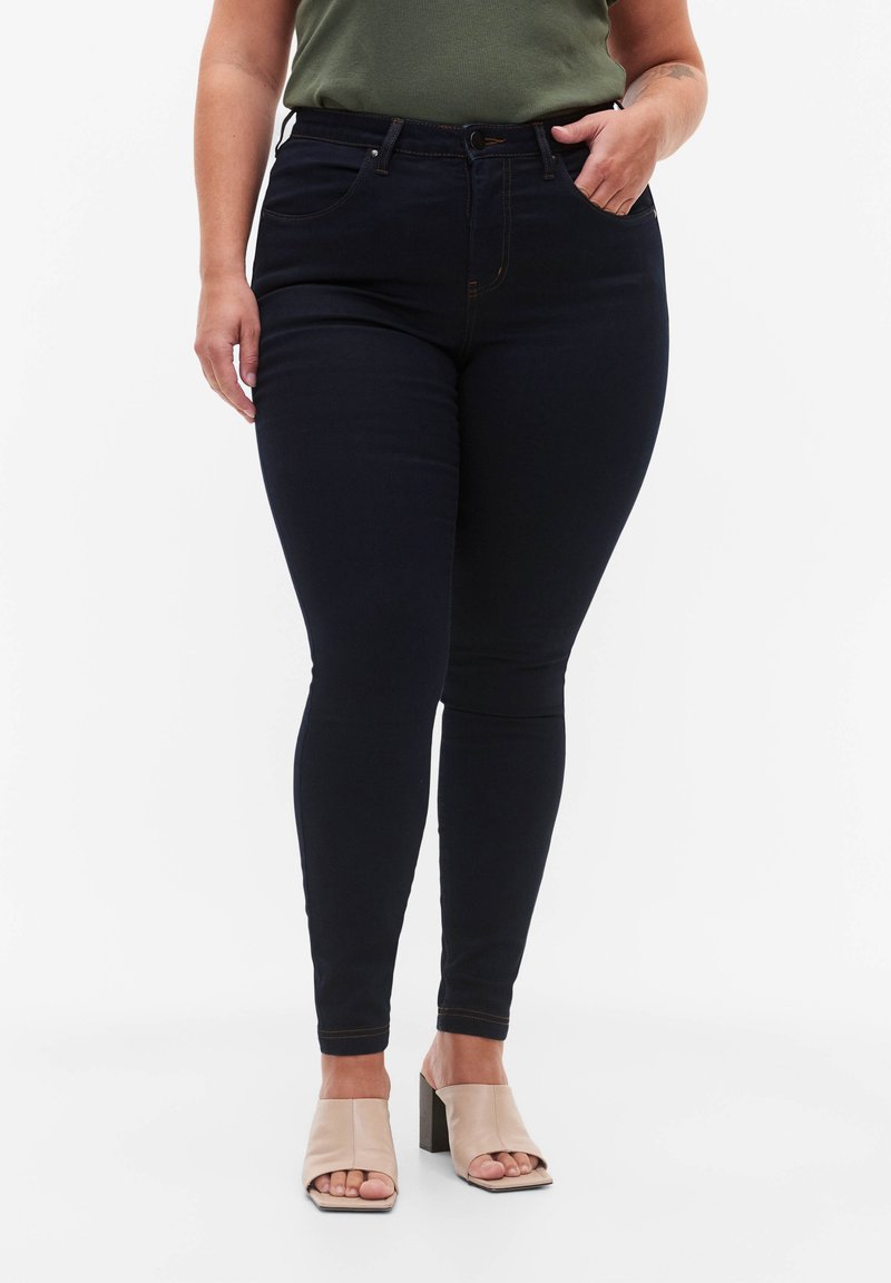 Vaqueros skinny de color azul oscuro con una cintura alta, ligera elasticidad, con bolsillos estándar y costuras doradas en contraste. Combinados con sandalias de tacón cuadrado beige.