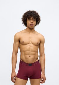 Boxer briefs de algodão bordeaux com um elástico preto largo rotulado "Calvin Klein." O modelo tem cabelo encaracolado e exibe um corpo superior tonificado.