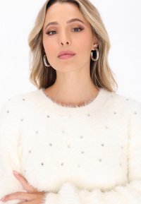 Blonde hajú nő ezüst karika fülbevalót visel, és egy fehér, bolyhos pulóvert, amelyet apró strasszok díszítenek.