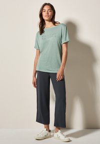T-shirt vert clair à manches courtes avec un motif graphique subtil, associé à un pantalon noir coupé et des baskets blanches avec des accents verts.