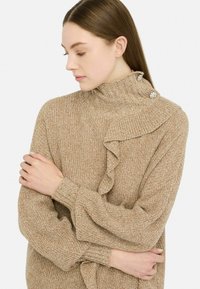 Maglione beige lavorato a maglia con colletto alto, caratterizzato da un design arricciato e tre bottoni decorativi sulla spalla. Maniche lunghe e a sbuffo.