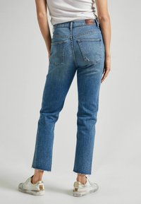 Osoba v modrých džínech s vysokým pasem a roztrhanými okraji, obutá v bílých teniskách se zlatými akcenty, zobrazená zezadu na neutrálním pozadí.