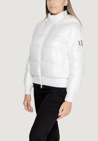 Giacca puffer bianca con colletto alto, tasche con zip e polsini a coste. Presenta un logo sulla manica e una superficie lucida e texturizzata.