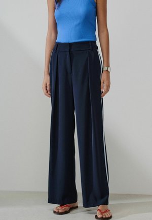 Femme portant un pantalon large bleu marine avec des rayures latérales blanches, un haut bleu sans manches, une montre et des sandales rouges string, debout devant un fond uni.