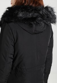 Černá prošívaná bunda s velkou, chlupatou černou umělou kožešinou na kapuci. Má přiléhavý střih a boční kapsy. Hladký povrch, zapínání na zip.