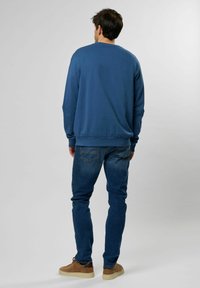 Felpa di cotone blu con collo rotondo, polsini a coste e orlo, abbinata a jeans in denim e sneakers marroni, vista da dietro.