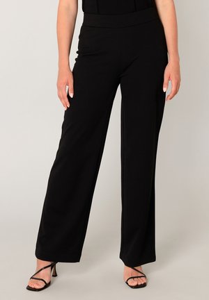 Vrouw die zwarte wijde broek met hoge taille draagt, gecombineerd met zwarte sandalen met bandjes en hak op een lichte achtergrond.