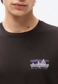 Κοντινό πλάνο ενός ατόμου που φοράει μαύρο πουκάμισο Patagonia με λευκό λογότυπο βουνού και το κείμενο «Save Our Home Planet» στο στήθος.