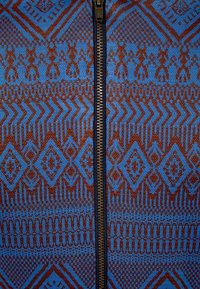 Vêtement tricoté zippé mettant en valeur des motifs géométriques complexes dans des nuances de bleu et de marron, avec une fermeture éclair en métal exposée au centre.