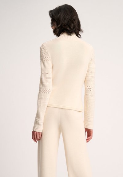 Cremfarbener, gestrickter Pullover mit hohem Kragen, der strukturierte Ärmel und einen taillierten Körper aufweist. Weicher Stoff mit unterschiedlichen Maschenmustern.